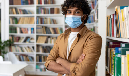 Portrait of young student inside library, man with crossed arms looking at camera, hispanic man with protective face mask visiting academic library and bookstore