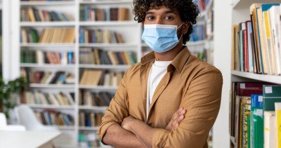 Portrait of young student inside library, man with crossed arms looking at camera, hispanic man with protective face mask visiting academic library and bookstore